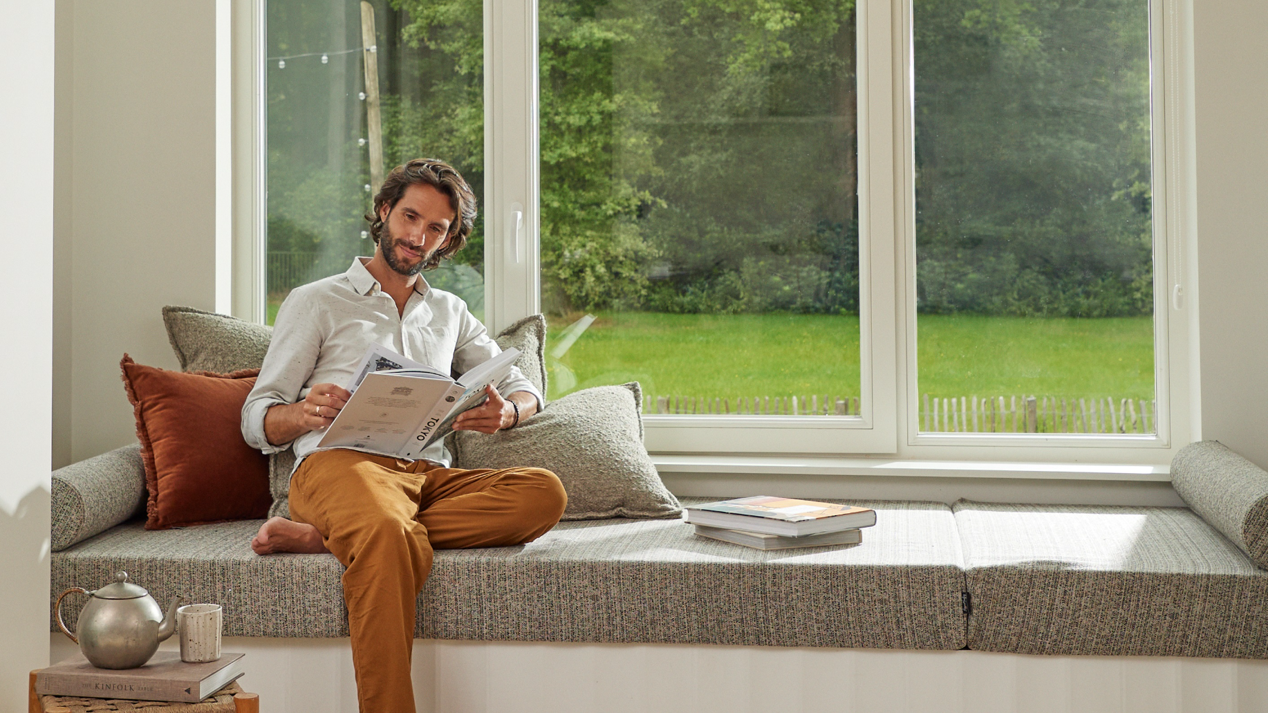 Ein Mann liest in einem gemütlichen Haus an einem großen Fenster und veranschaulicht den Komfort und die Energieeffizienz von Wärmepumpen bei der Verringerung der Umweltauswirkungen von Heizungslösungen für Innenräume.