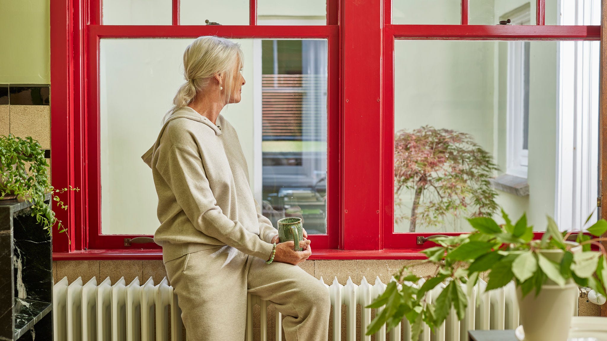 Frau, die an einem Heizkörper neben einem großen Fenster sitzt und ein warmes, energieeffizientes Zuhause genießt