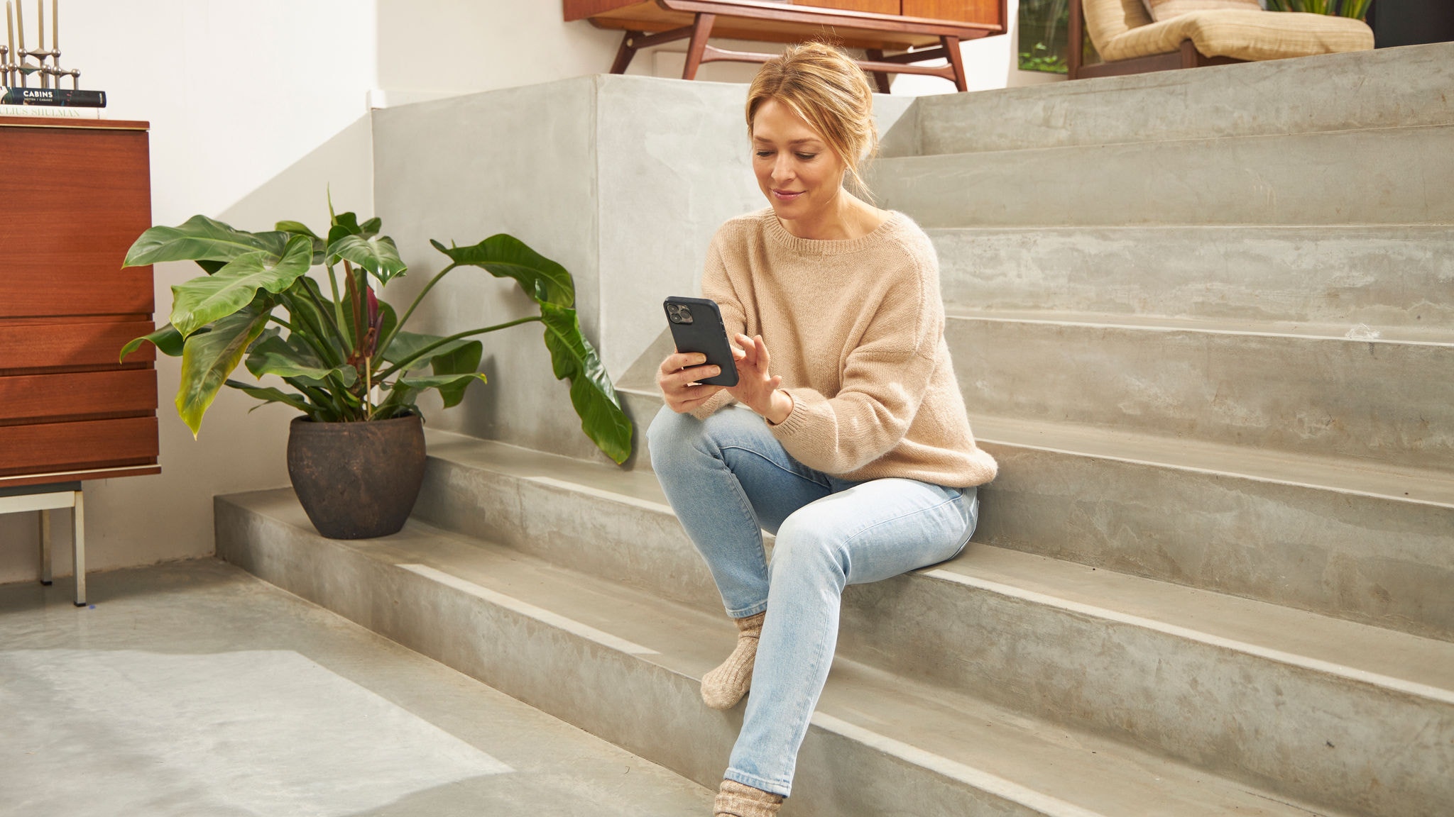 Eine Frau sitzt Zuhause auf der Treppe und bedient die Onecta App auf ihrem Smartphone.