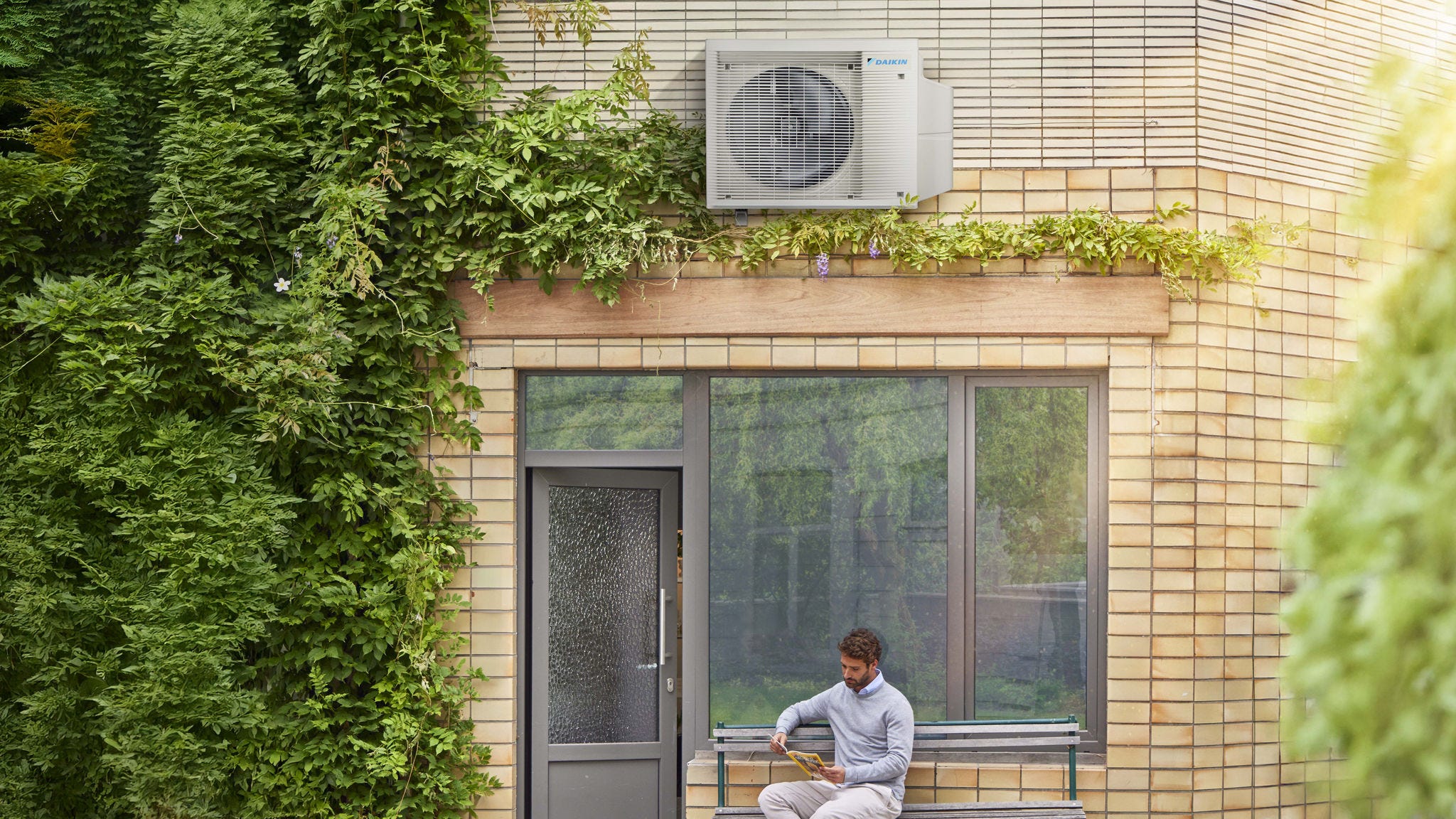 Ein Mann liest auf einer Bank vor seinem Haus unter einer DAIKIN Luft-Wärmepumpe.