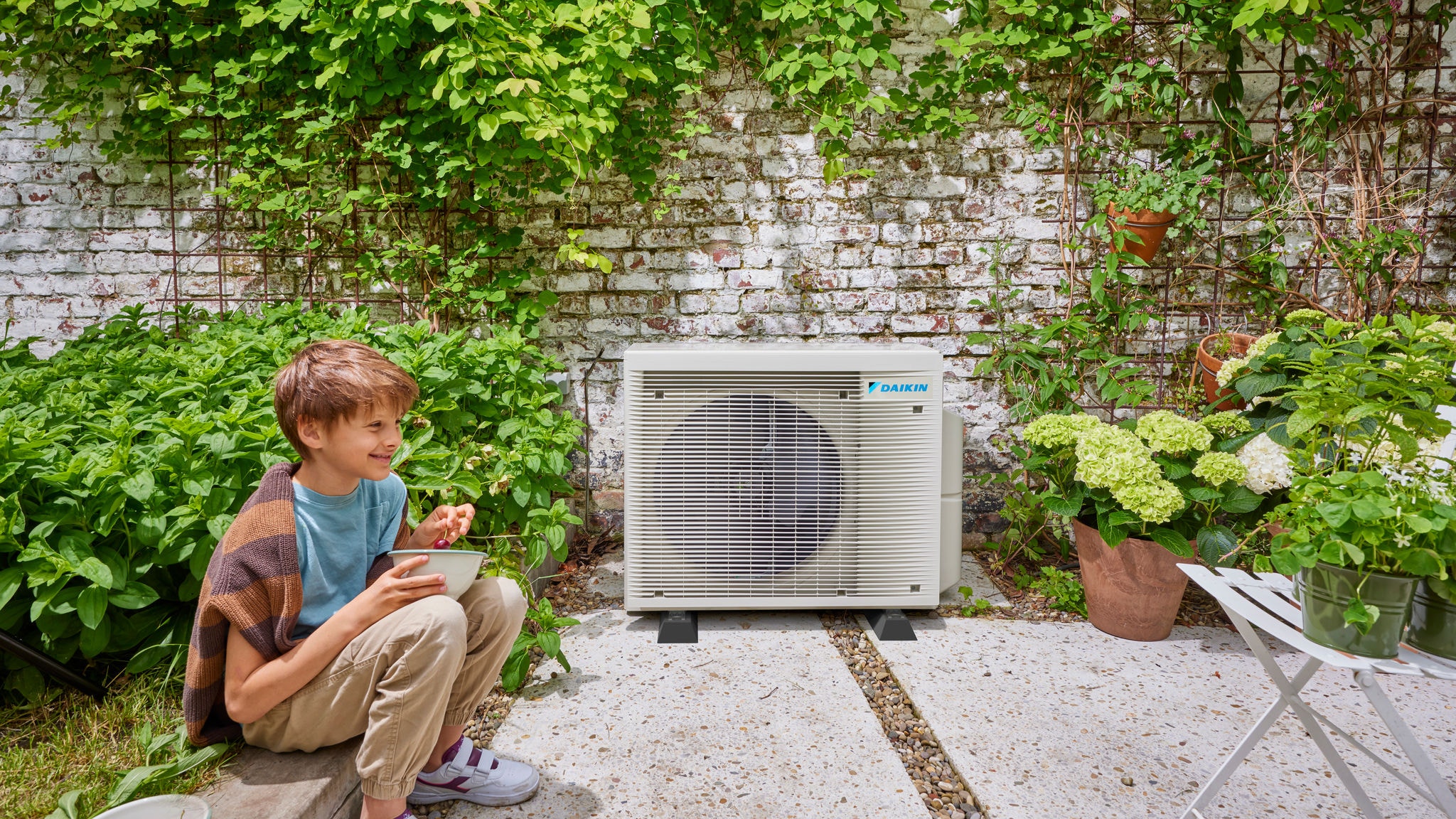 Ein Kind neben einem Außengerät einer DAIKIN Hybrid-Wärmepumpe, das Energieeffizienz und einen netten Garten vereint