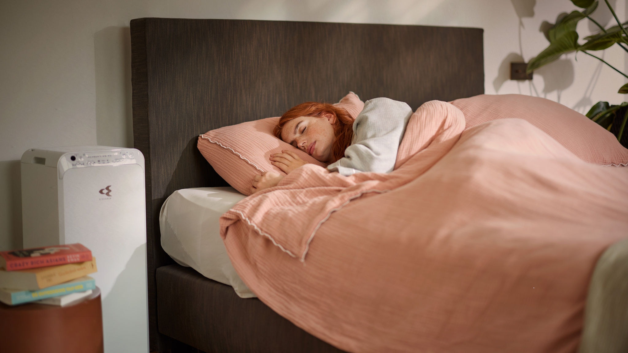 Eine Frau schläft friedlich in einem kuscheligen Bett, während in der Nähe ein Luftreiniger arbeitet, der im Winter im Schlafzimmer für saubere Luft sorgt