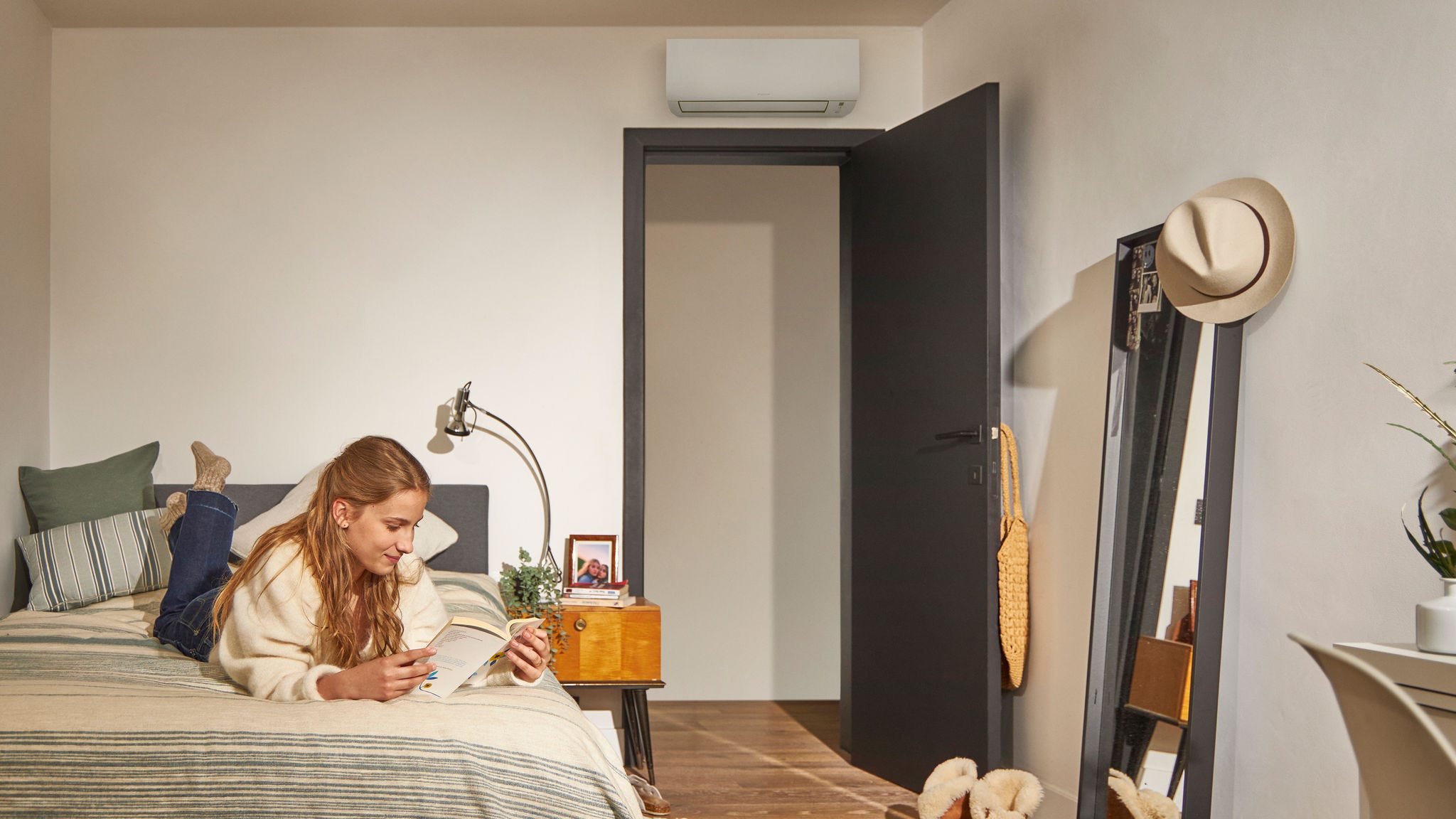 Eine Frau liest auf einem Bett in einem gemütlichen Schlafzimmer; eine Wärmepumpen-Klimaanlage über der Tür sorgt für eine energieeffiziente Klimatisierung