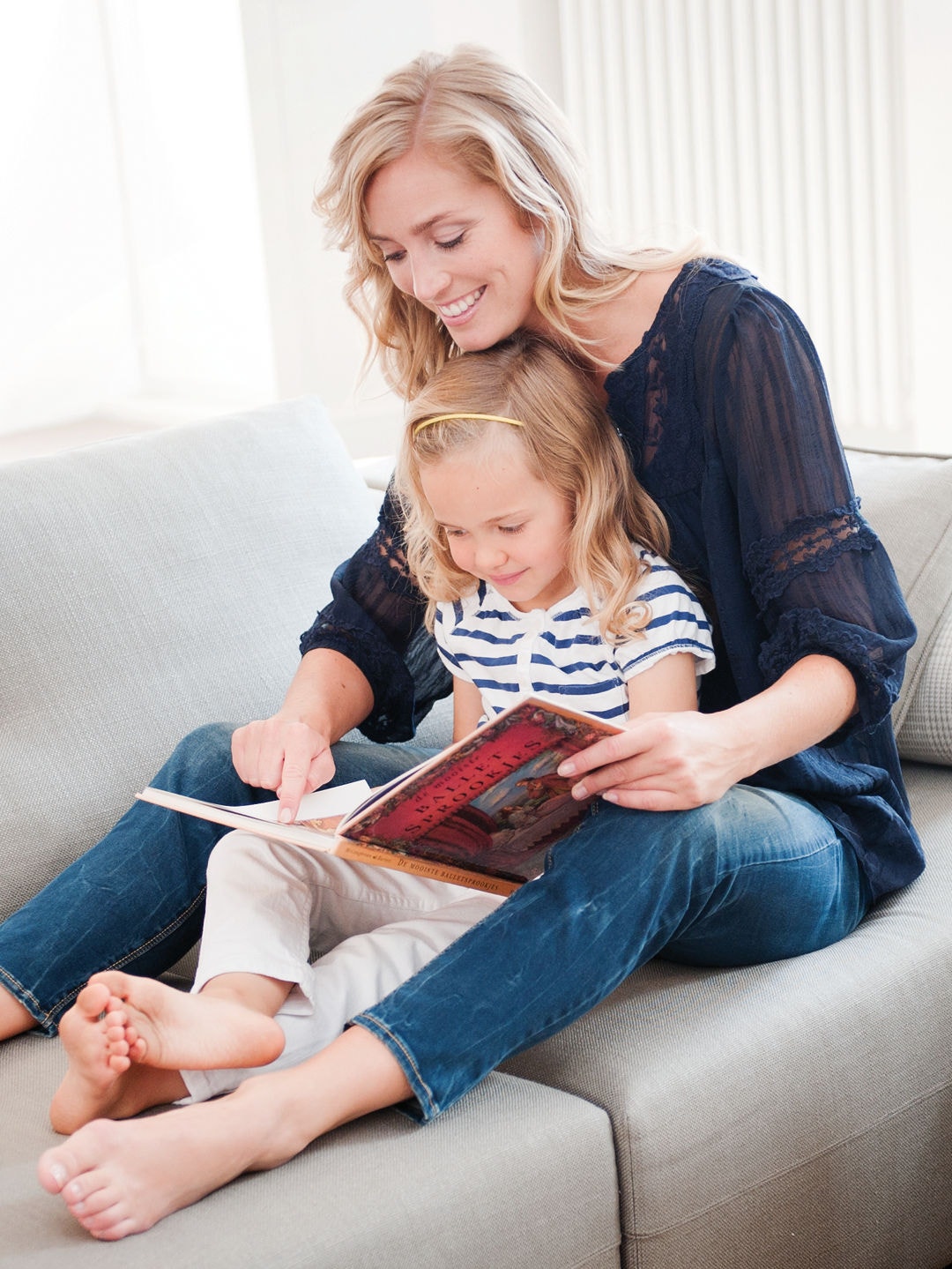 Kleines Mädchen sitzt auf dem Schoß ihrer Mutter auf dem Sofa, und beide lesen gemeinsam ein Buch bei perfekter Raumtemperatur