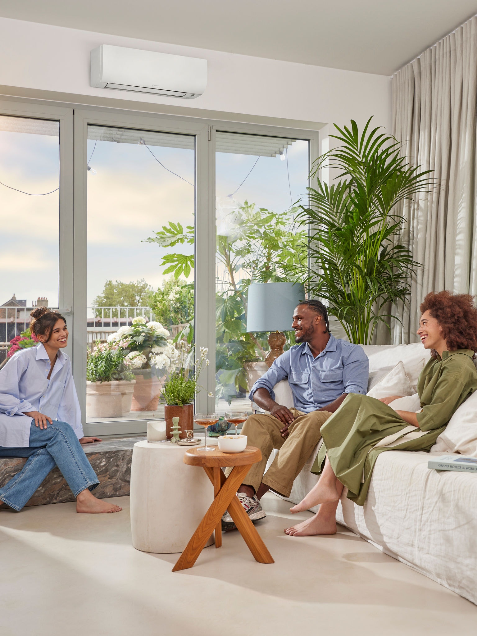 Ein Mann und zwei Frauen sitzen an einem sonnigen Tag in einem hellen, luftigen Raum mit weißen Wänden und unterhalten sich. Über einem großen Bild ist ein unauffälliges, weißes Wärmepumpen-Wandgerät zu sehen