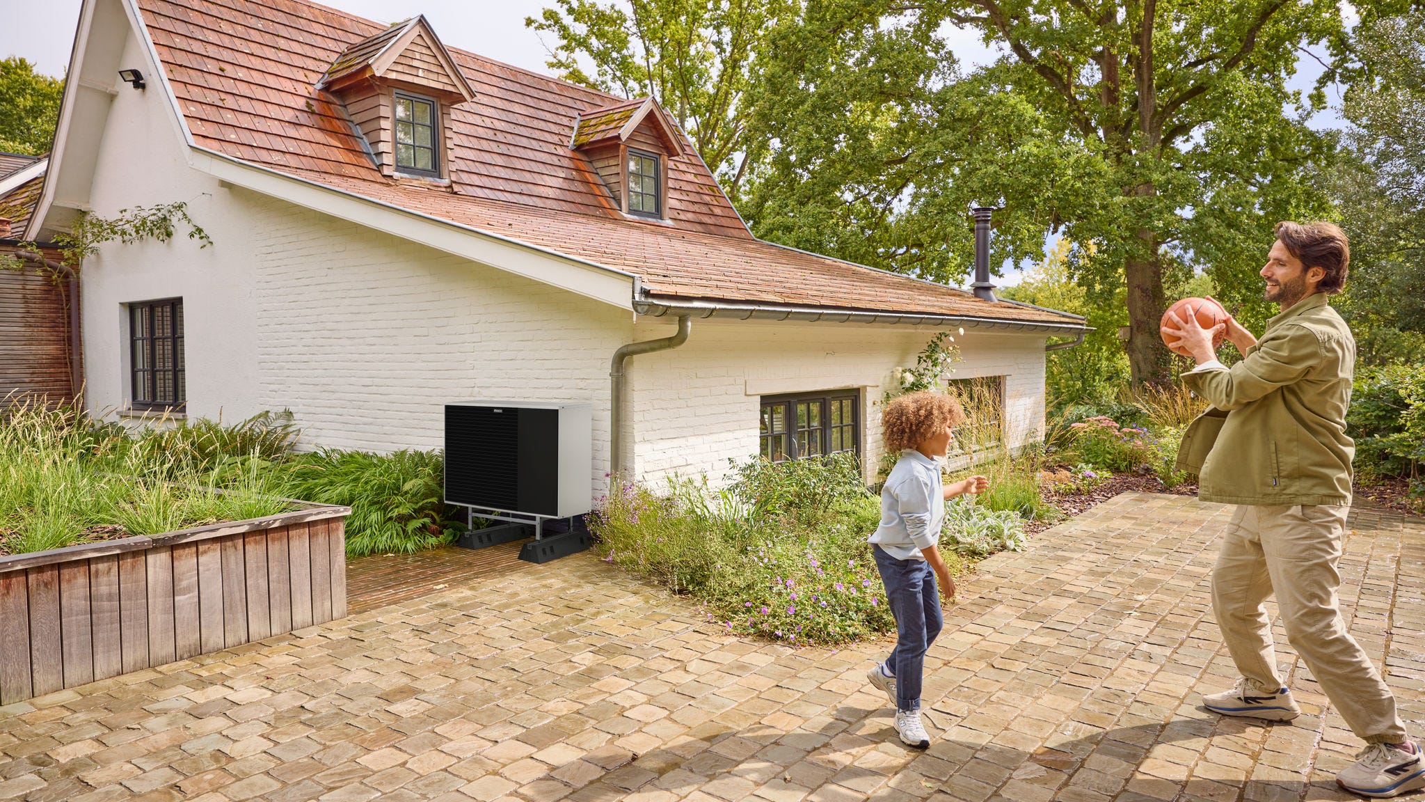 Vater und Kind spielen im Freien Ball auf dem Hof, auf dem eine Wärmepumpe installiert ist
