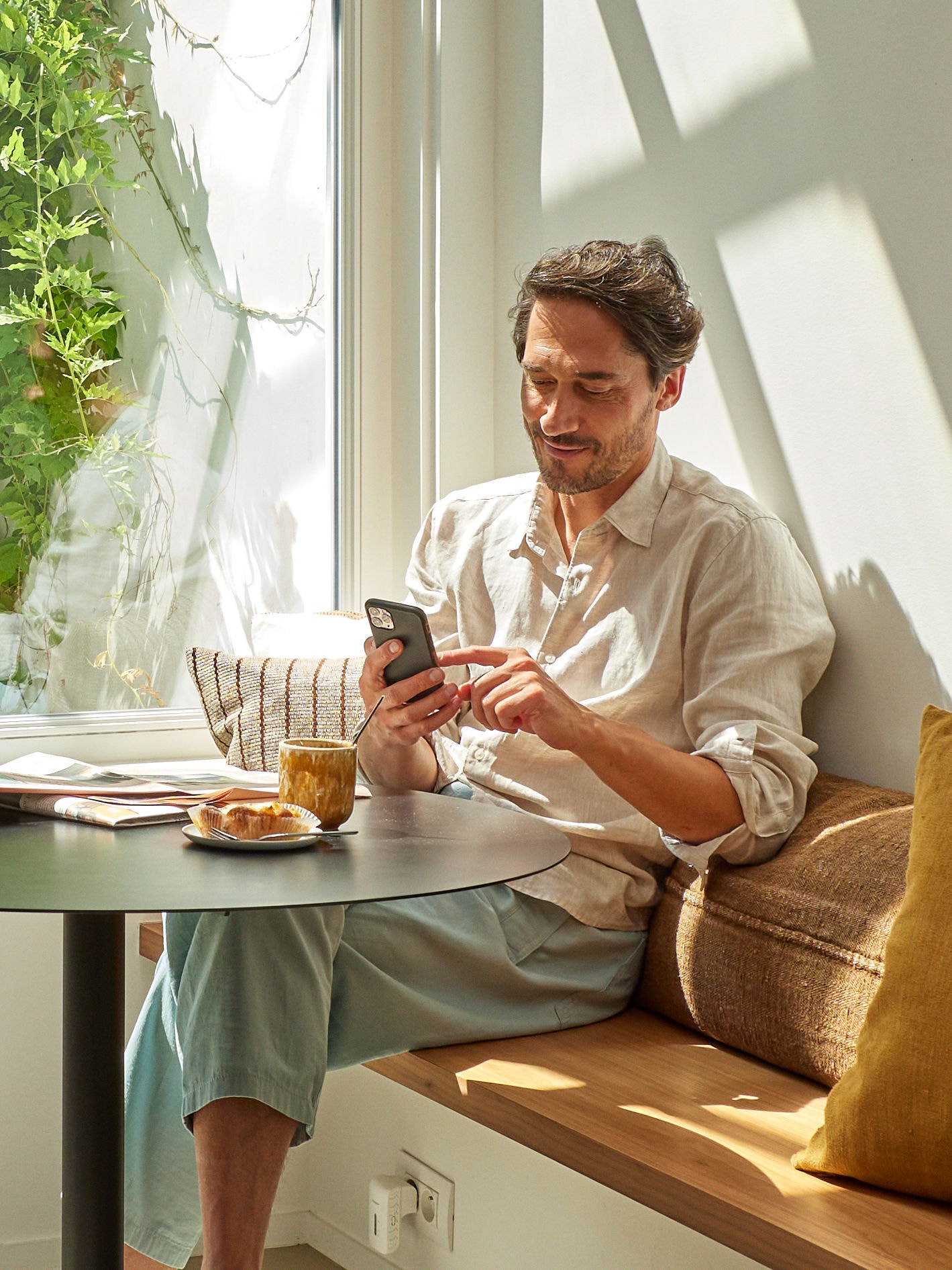 Mann sitzt mit einem Kaffee und Imbiss entspannt am Tisch und schaut auf sein Smartphone