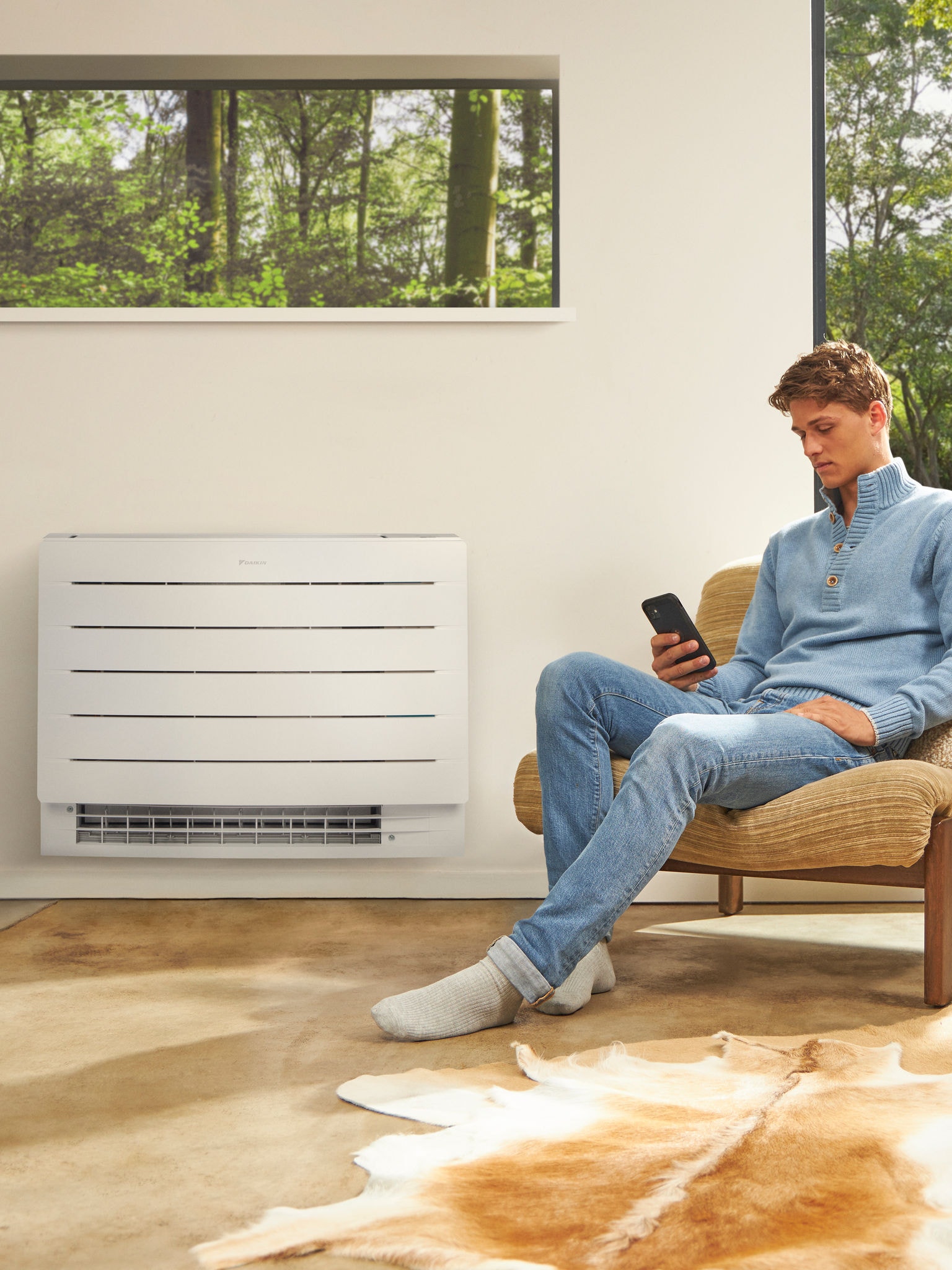 Ein Mann sitzt in einem hellen Raum und schaut auf sein Telefon, an der Wand hinter ihm ist eine Wärmepumpe