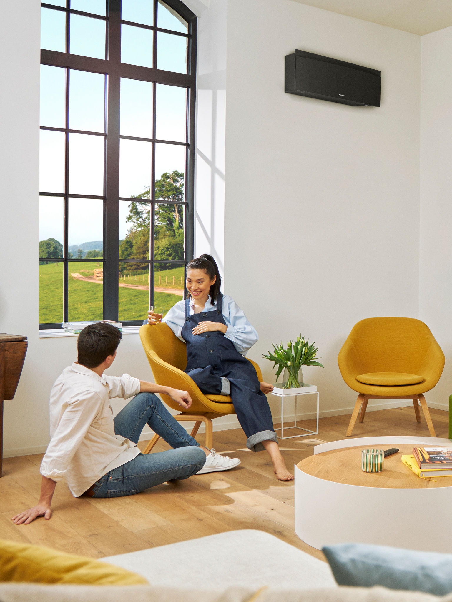 Ein Mann und eine Frau sitzen vor einem großen Fenster in einem hellen Raum. Hinter ihnen an der Wand ein schwarzes Wärmepumpen-Wandgerät