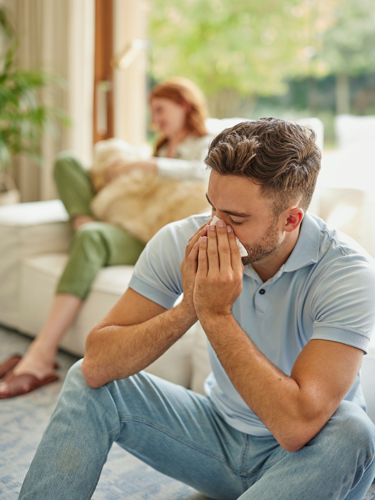 Eine Frau im Hintergrund auf Polstermöbeln mit ihrem Golden Retriever auf dem Schoß, ein Mann im Vordergrund niest